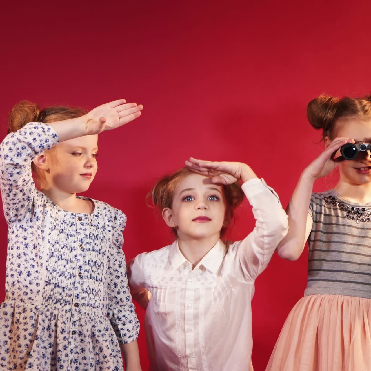 three young girlfriend are considering everything around themselves on the red background. One of little girl in dress is looking through binoculars