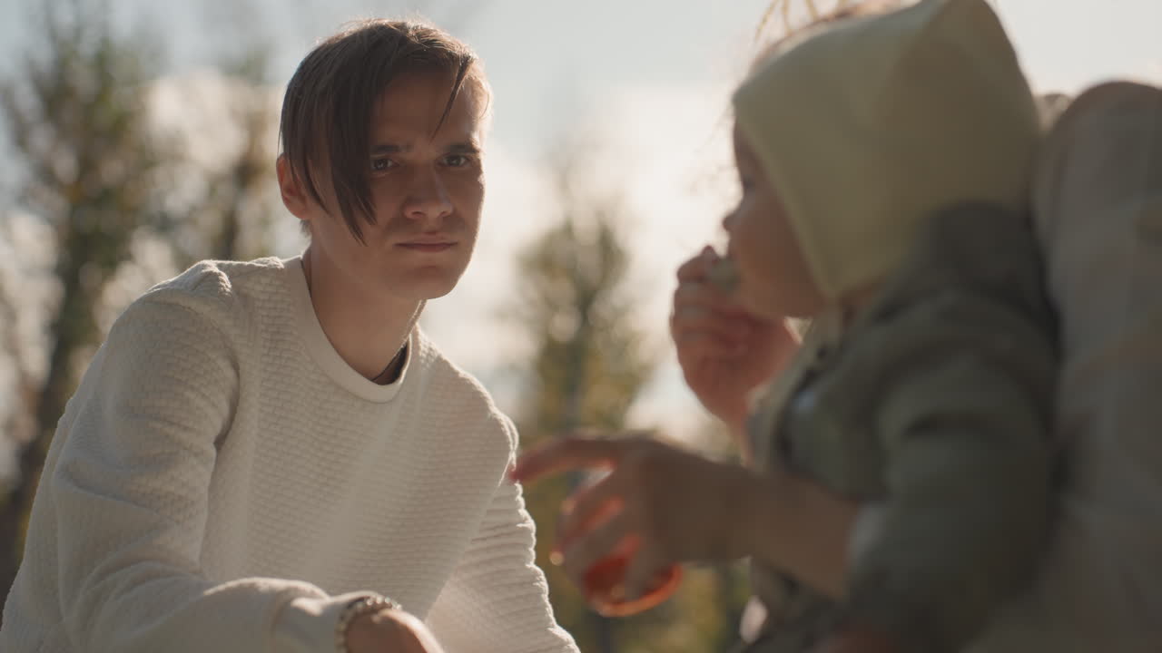Forelder mater smårolling utendørs i sollys, ung voksen i hvit genser ser ømt på barn i strikkelue, deler snacks med skje og kopp, myk høsthagebokeh, varm koselig stemning, subtil familie