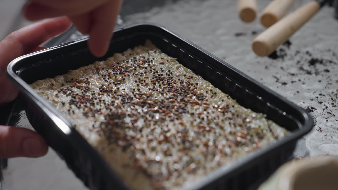 Urban gardener hands gently spreading tiny seeds from ceramic pot onto moist textured growing medium in black tray on glass table with scattered tools and green plants showcasing serene indoor sowing