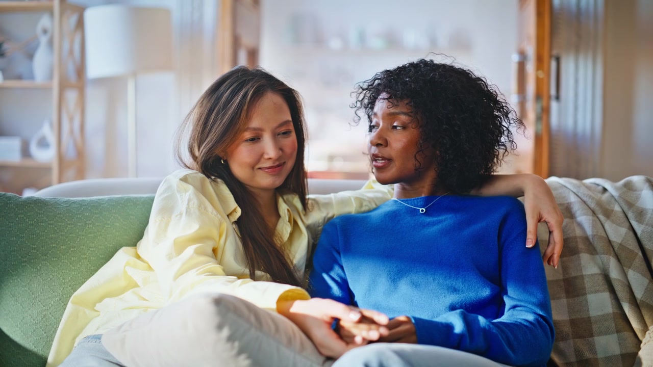 Pareja de lesbianas acurrucándose en el sofá de casa. Mujeres interraciales abrazándose en la sala de estar