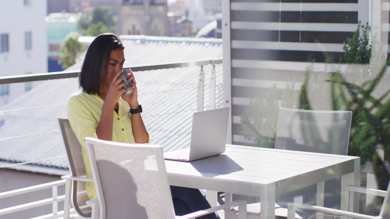 Mixed race gender fluid person drinking cup of coffee and using laptop on roof terrace