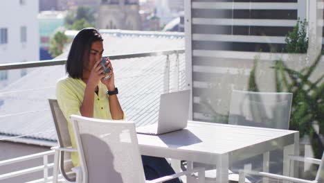 mixed race gender fluid person drinking cup of coffee and using laptop on roof terrace