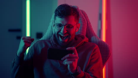 A-young-man-with-glasses-a-professional-gamer-on-a-mobile-phone-plays-games-and-emotionally-rejoices-in-the-victory.-Emotional-young-man-playing-mobile-games-in-a-neon-light.-High-quality-4k-footage