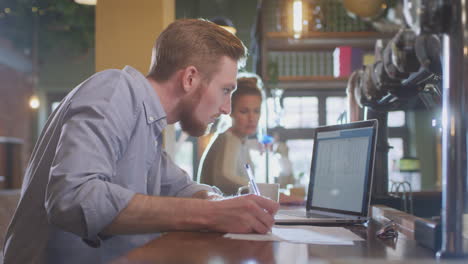 Male-Manager-Of-Bar-Working-At-Laptop-On-Counter-Doing-Accounts-With-Woman-Cleaning-In-Background