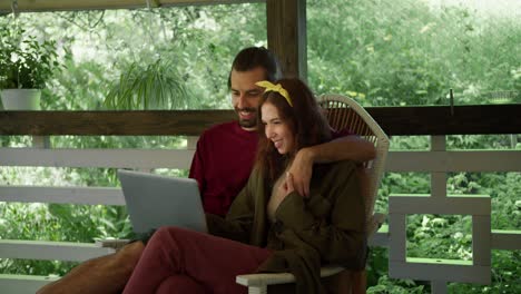 A-brunette-guy-in-a-red-blouse-and-a-brunette-girl-in-a-green-blouse-are-sitting-on-an-armchair,-talking-and-looking-at-something-in-a-laptop-in-the-gazebo-against-the-backdrop-of-a-green-forest