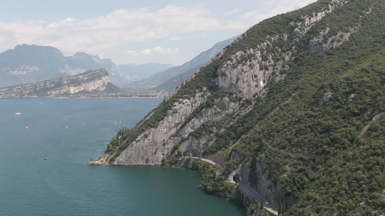 Ripresa aerea del Lago di Garda con piccole barche a vela sul lago, sullo sfondo montagne e sulla destra auto su una strada in una giornata di sole.