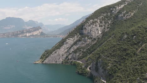 Drone-shot-of-Lake-Garda-with-small-sailing-boats-on-the-lake-in-the-background-are-mountains,-on-the-right-are-cars-on-a-road-on-a-sunny-day