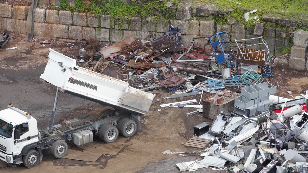 A dump truck unloads metal scrap in a junkyard, showcasing industrial recycling processes. The environment is gritty with overcast lighting
