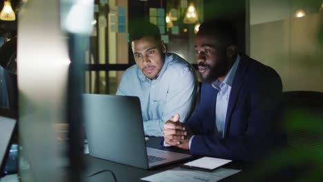 Video-of-two-happy-diverse-businessmen-talking-and-using-laptop-together-at-night-in-office