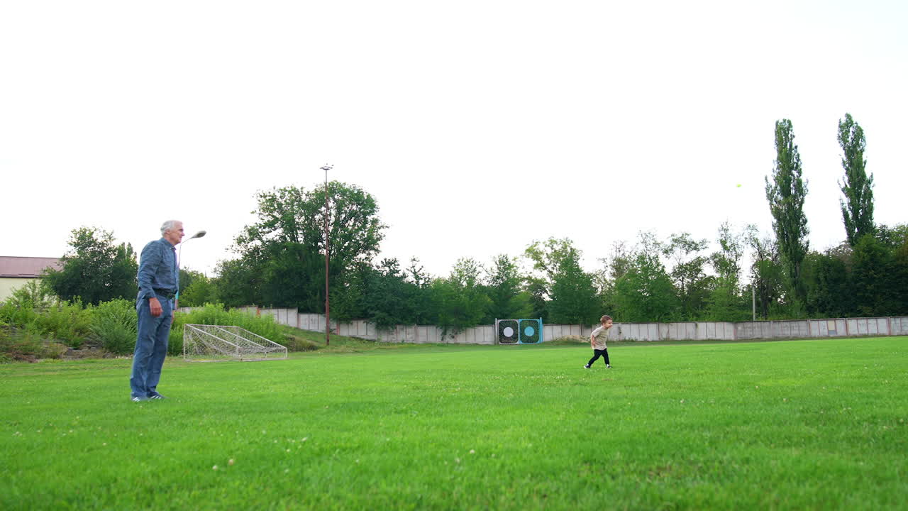Senior man standing on the green field tosses a little ball. Small kid watches the ball fly. Grandpa and grandson play outdoors in summer.