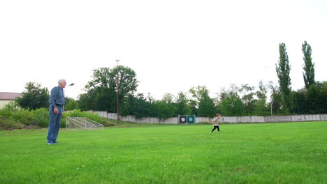 senior man standing on the green field tosses a little ball. small kid watches the ball fly. grandpa and grandson play outdoors in summer.
