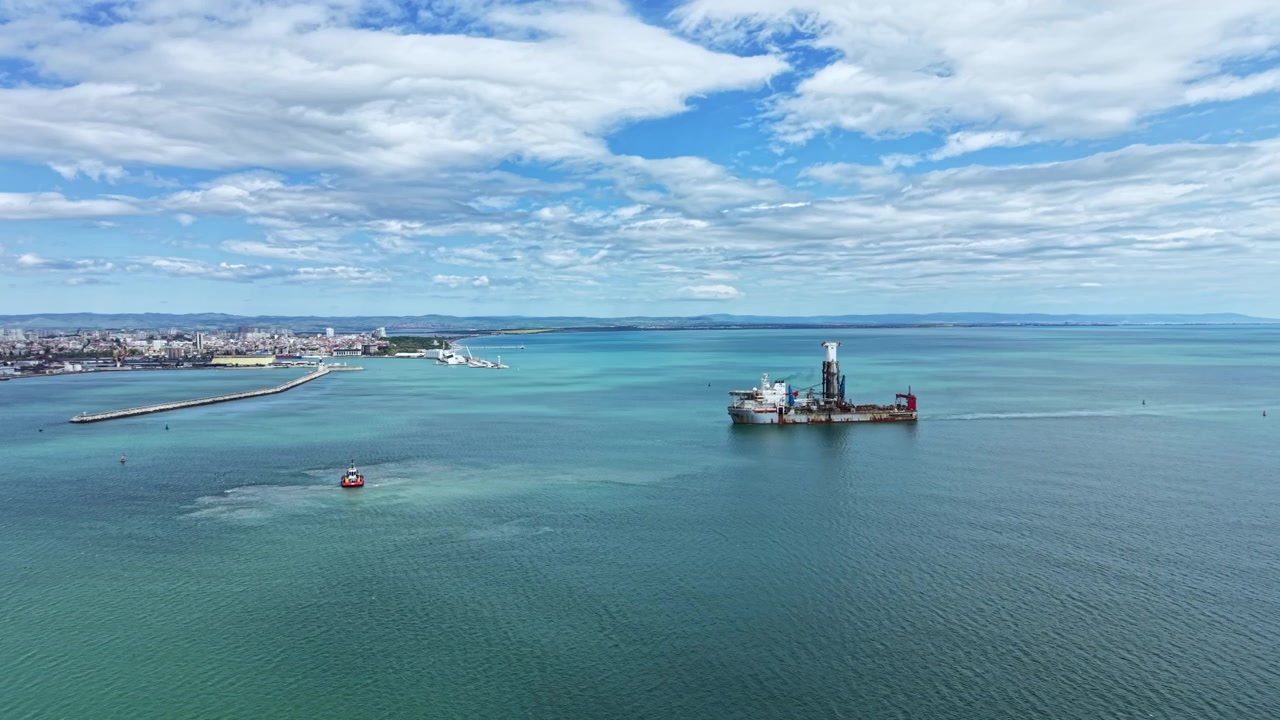 Industrial dredging vessel works near coastline by a port under a clear sky with some clouds during daytime