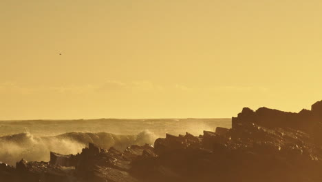 Raue-Meereswellen-Brechen-Gegen-Felsen-Und-Erzeugen-Bei-Goldenem-Sonnenuntergang-Eine-Riesige-Gischt---Mittlere-Schwenkaufnahme