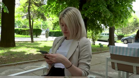 beautiful blonde girl sitting at a table, texting on her phone and checking surroundings, outside, summer, vacation