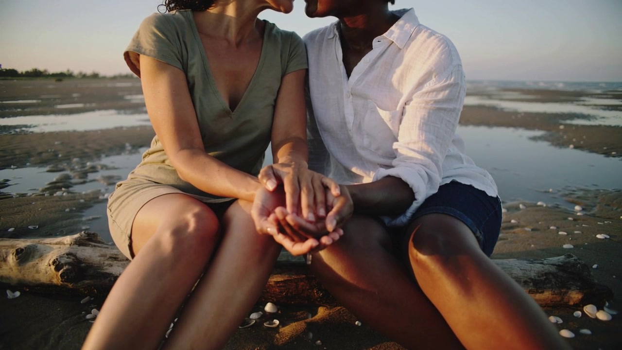 Di fronte al tramonto, una coppia in abito e camicia si appoggia, tenendo le mani a coppa su un pezzo di legno portato dalla corrente in spiaggia.