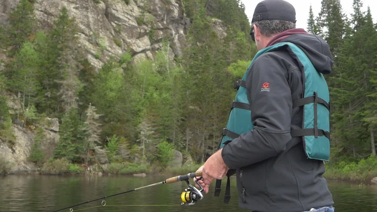 Slow-motion medium shot of fisherman reeling in fishing line with lake and water in background.