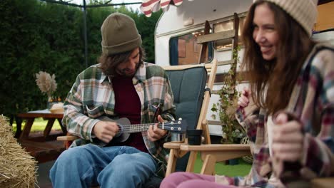 a happy brunette guy in a green hat in a green checkered shirt plays a black ukulele and his brunette girlfriend in a pink checkered shirt is a trailer dancer during their picnic in a camp outside the city in the summer