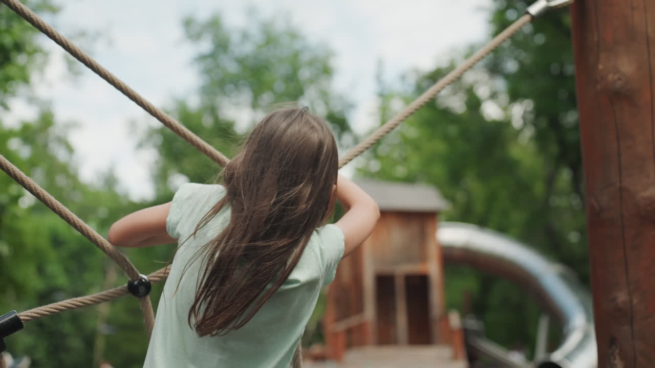 Daughter climbing rope net in park, focused grip on woven ropes and wooden posts, steady balance and careful footwork, green trees backdrop, summer outdoor challenge, playful training and exploration