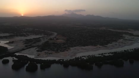 drone moves towards the horizon with revealing a river and mangrove forest, riverbank, highway on the lefthand side, progressing sunset behind the mountains, porlamar, margarita island, venezuela