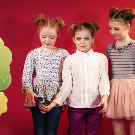 three happy girls playing together with binoculars on the red background. pretty little girls looking into the black binoculars in childish studio with trees decor.