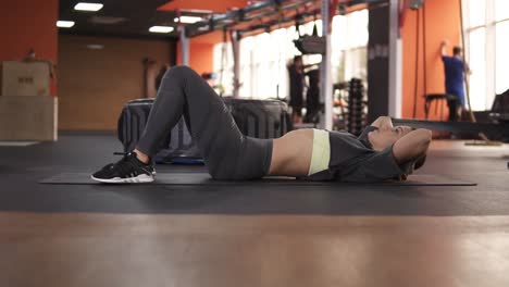 Young-european-woman-in-grey-sportswear-pumps-the-abdominal-muscles-on-the-floor-in-the-modern-gym.-Sport-and-fitness-concept.-Footage-from-the-side.-Slow-motion