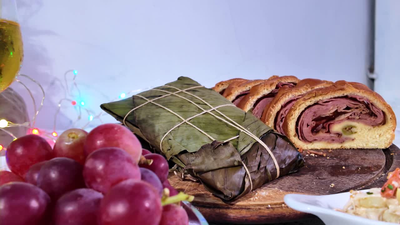 tracking shot showcasing a festive table with traditional Venezuelan holiday dishes: a cornmeal Hallaca wrapped in banana leaves and sliced Pan de Jamon (ham bread), served with grapes, nuts, and wine