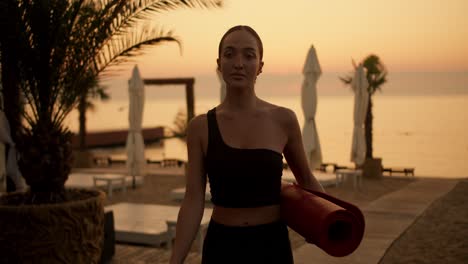 Una-Chica-De-Mediana-Edad-Con-Un-Chándal-Negro-Y-Una-Estera-Roja-En-Las-Manos-Saliendo-De-La-Playa-Después-De-Una-Clase-De-Yoga-Y-Ejercicio-Nocturno.-Hora-Dorada,-Atardecer-Dorado-En-La-Playa