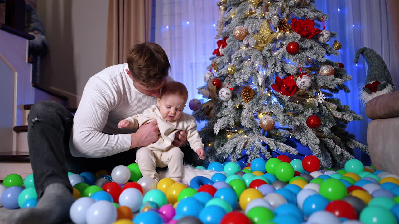 Father and son play with the plastic ball scattered on the floor near the Christmas tree. Loving dad kisses his son on the head.