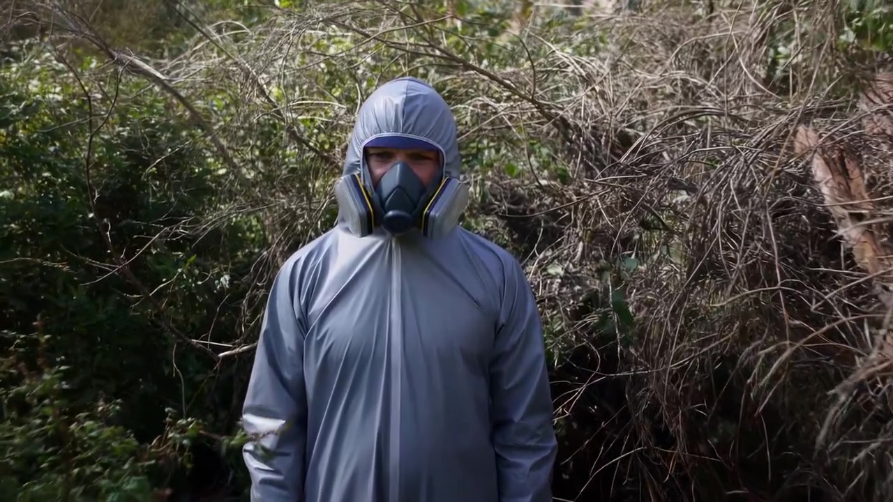 Uma pessoa com um traje de proteção está parada em uma área arborizada ao ar livre, demonstrando medidas de segurança e resposta a emergências. O ambiente neutro e os tons cinza-esverdeados criam uma atmosfera profissional e calma.