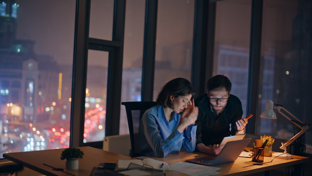 Tensed coworkers analyzing problem online project at night office. Two partners