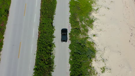 SUV-driving-on-empty-white-sand-beach-coastline-road,-aerial-top-down