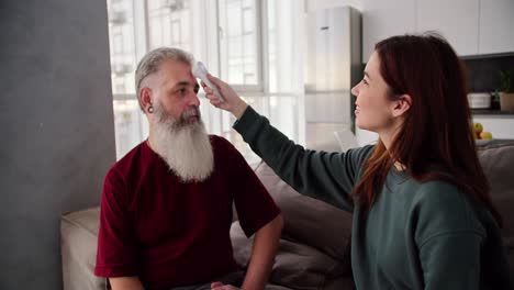 A-brunette-girl-in-a-Green-jacket-measures-the-temperature-with-an-electronic-thermometer-by-applying-it-to-the-forehead-of-her-elderly-father-with-gray-hair-and-a-lush-beard-in-a-red-T-shirt-on-a-modern-sofa-in-the-apartment