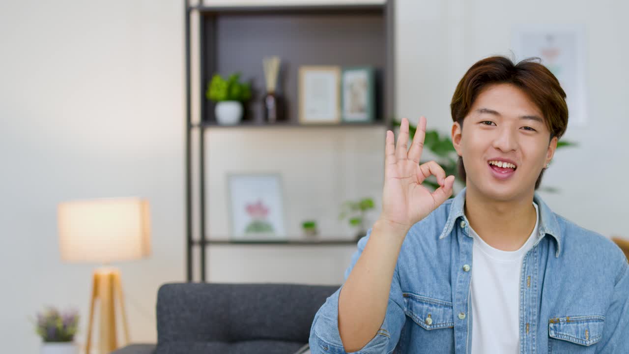 Uomo asiatico sorridente con una camicia di jeans che fa un segno di okay in un salotto