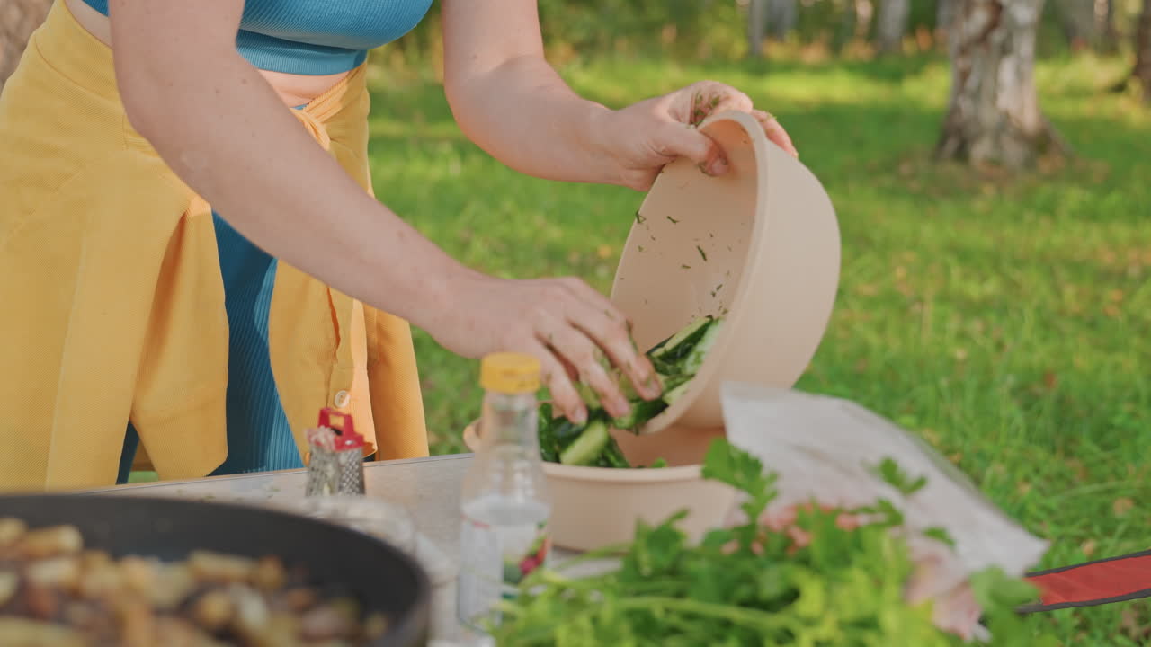 Mano che mescola un'insalata da campeggio in una ciotola. Una donna versa verdure da una grande ciotola, aggiungendo erbe aromatiche. Una bottiglia di condimento è vicina, una padella con funghi in primo piano, un tavolo di legno, uno sfondo di betulle, luce diurna intensa.
