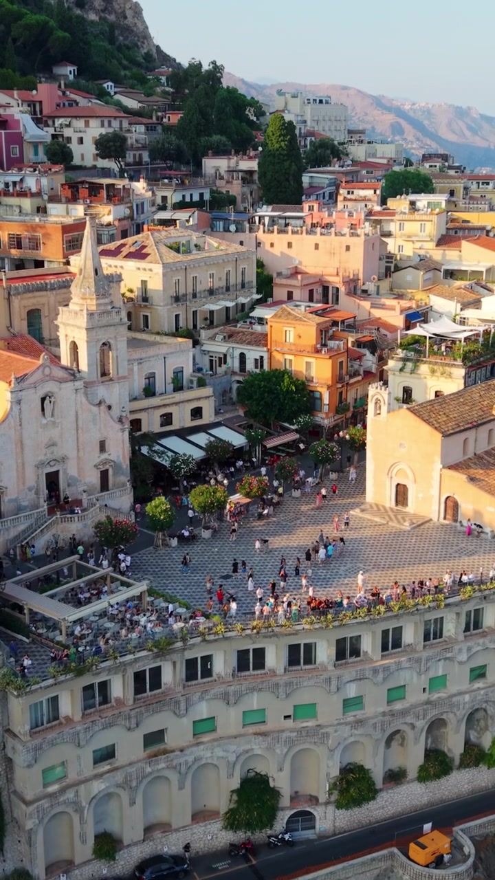 Veduta aerea di una vivace piazza cittadina con edifici storici a Taormina in una giornata di sole.
