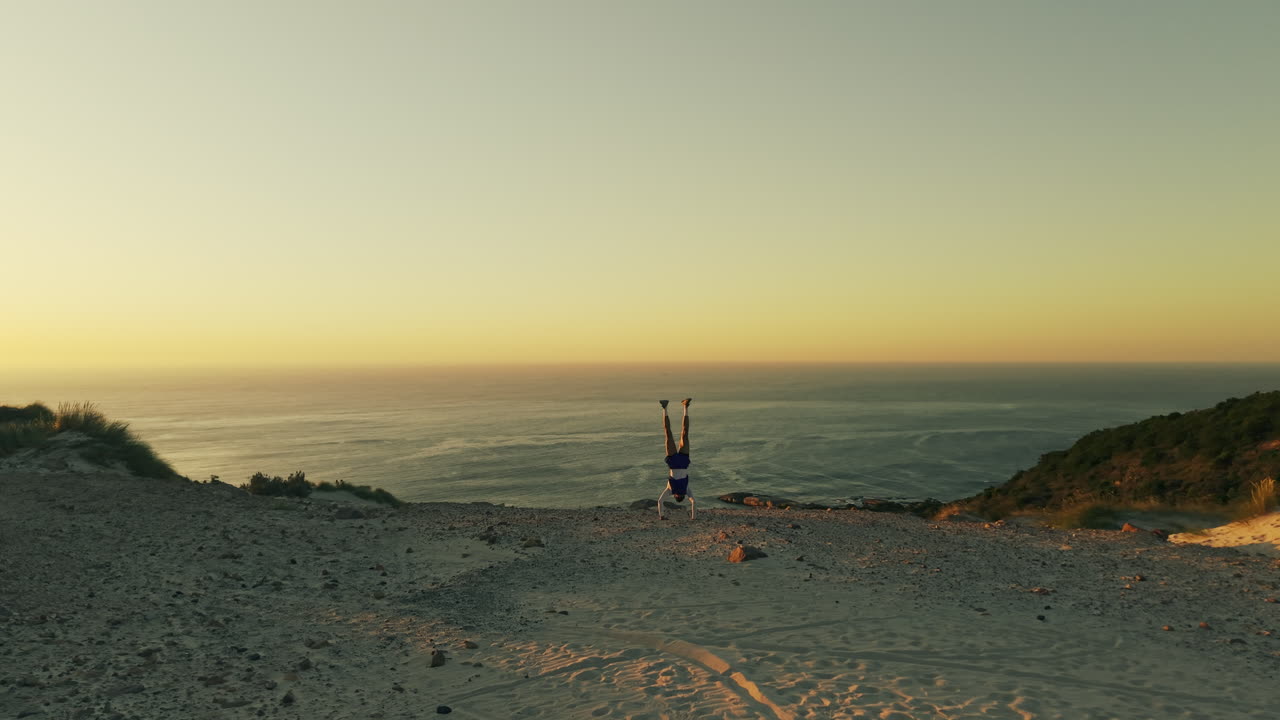 Persona che fa la verticale su una scogliera sabbiosa con vista sull'oceano al tramonto