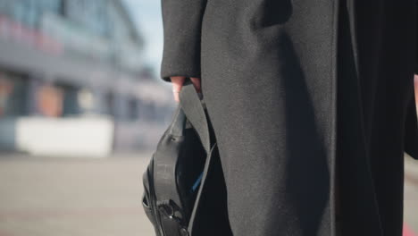 close-up of a black bag held by a person wearing a stylish black coat and leather boots, with natural sunlight highlighting the elegant details of the outfit on an urban street backdrop