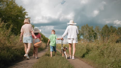 parent and children walking along dirt road surrounded by lush greenery going for picnic, carrying basket while dog walks on leash under partly cloudy sky