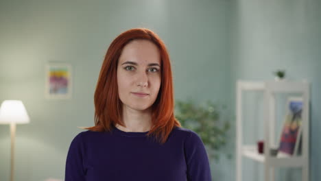 Young-redhead-woman-looks-in-camera-slightly-smiling-in-room