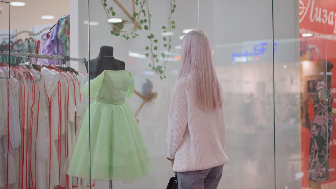 young buyer with bag approaches glass display to admire short green flare gown on mannequin, boutique lights reflecting on shimmering tulle skirt and bodice details creating elegant mood
