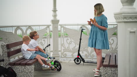 a mother in a blue gown interacts with her two children while holding pastries in her hand, one child sits next to a scooter while the mother steps slightly forward