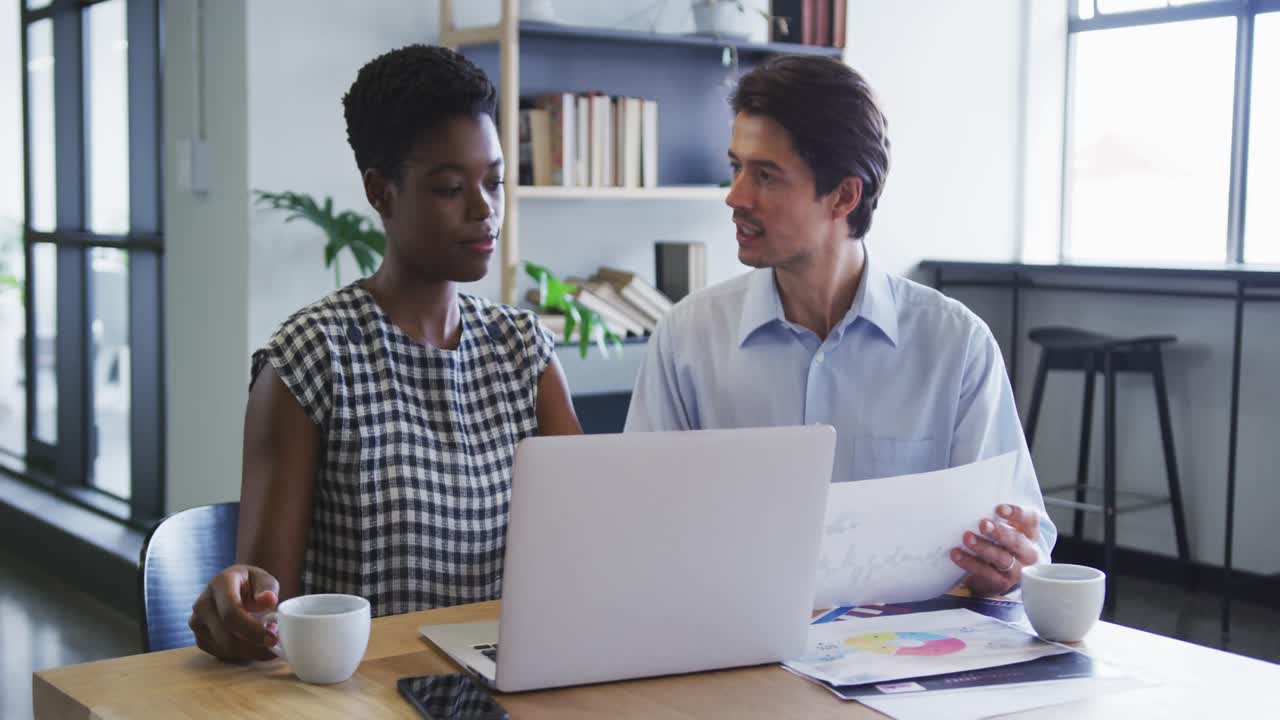 Diverse business colleagues sitting at desk using laptop going through paperwork in office