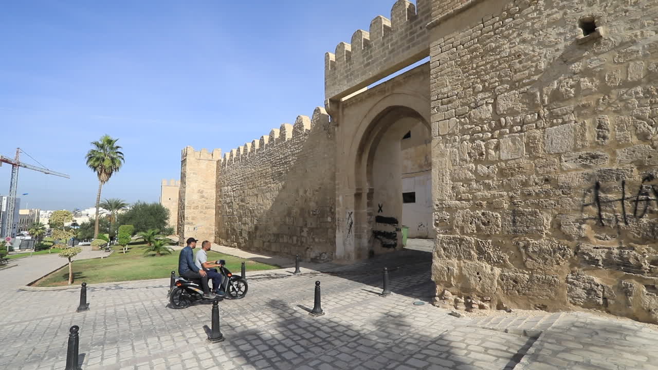 Two people ride a motorcycle past the ancient city walls under a clear blue sky on a sunny day