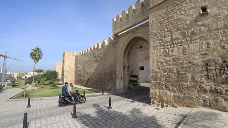 two people ride a motorcycle past the ancient city walls under a clear blue sky on a sunny day