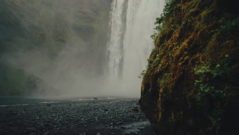 reveal shot from behind cliffs of scenic misty skógafoss falls in iceland, slomo