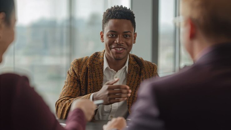 Reaching man in brown blazer responding to document handed from right in office, wristwatch visible
