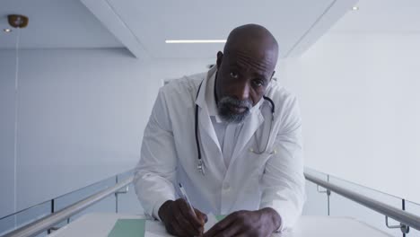 Portrait-of-african-american-male-senior-doctor-taking-notes-while-looking-at-the-camera-at-hospital