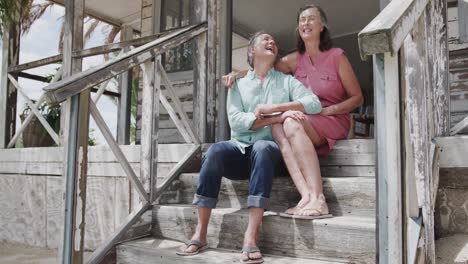 Happy-senior-caucasian-couple-embracing-and-talking-sitting-on-steps-of-beach-house,-in-slow-motion