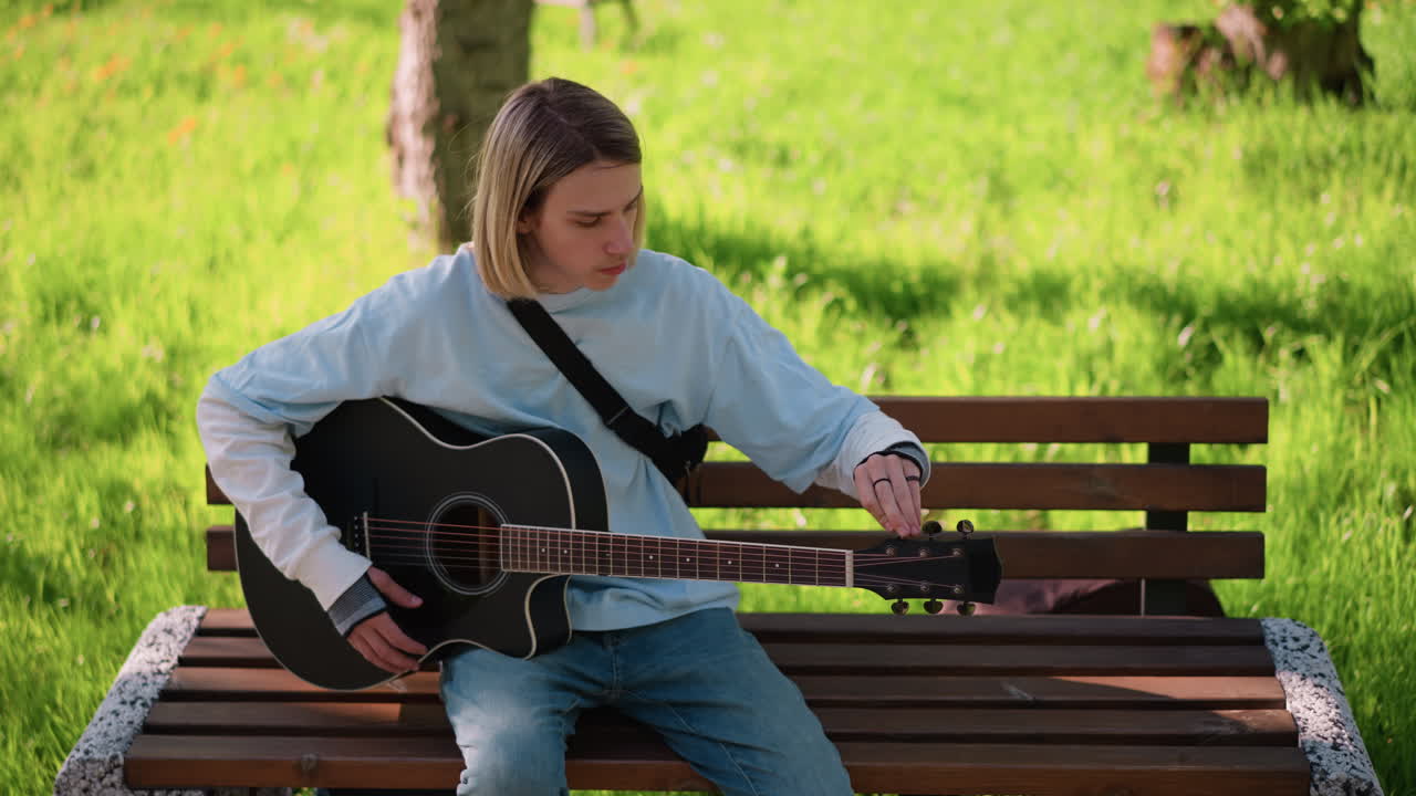 chitarrista su una panchina illuminata dal sole che suona la chitarra, sessione rilassata di pratica e composizione di canzoni su una panchina di legno, felpa casual e denim, dolce strumming, alberi verdi ed erba sullo sfondo, concentrato