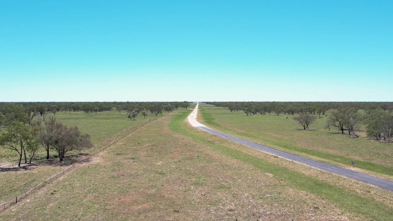Vuelo de dron a baja altitud siguiendo un estrecho camino rural a través de tierras de cultivo abiertas con árboles dispersos y terreno seco, creando una perspectiva de movimiento hacia adelante y una escena de viaje de larga distancia bajo un cielo despejado.